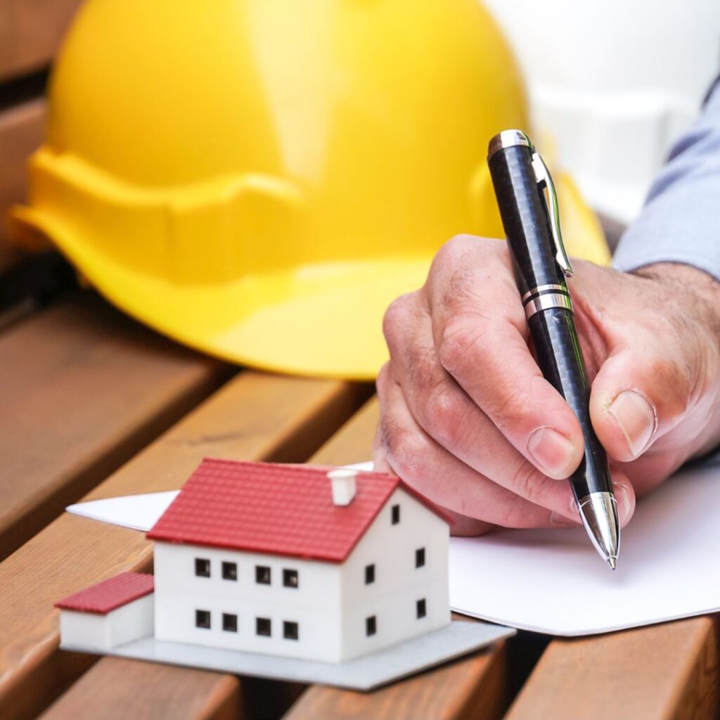 Image of a hand signing a paper with a helmet near and a small model of a house
